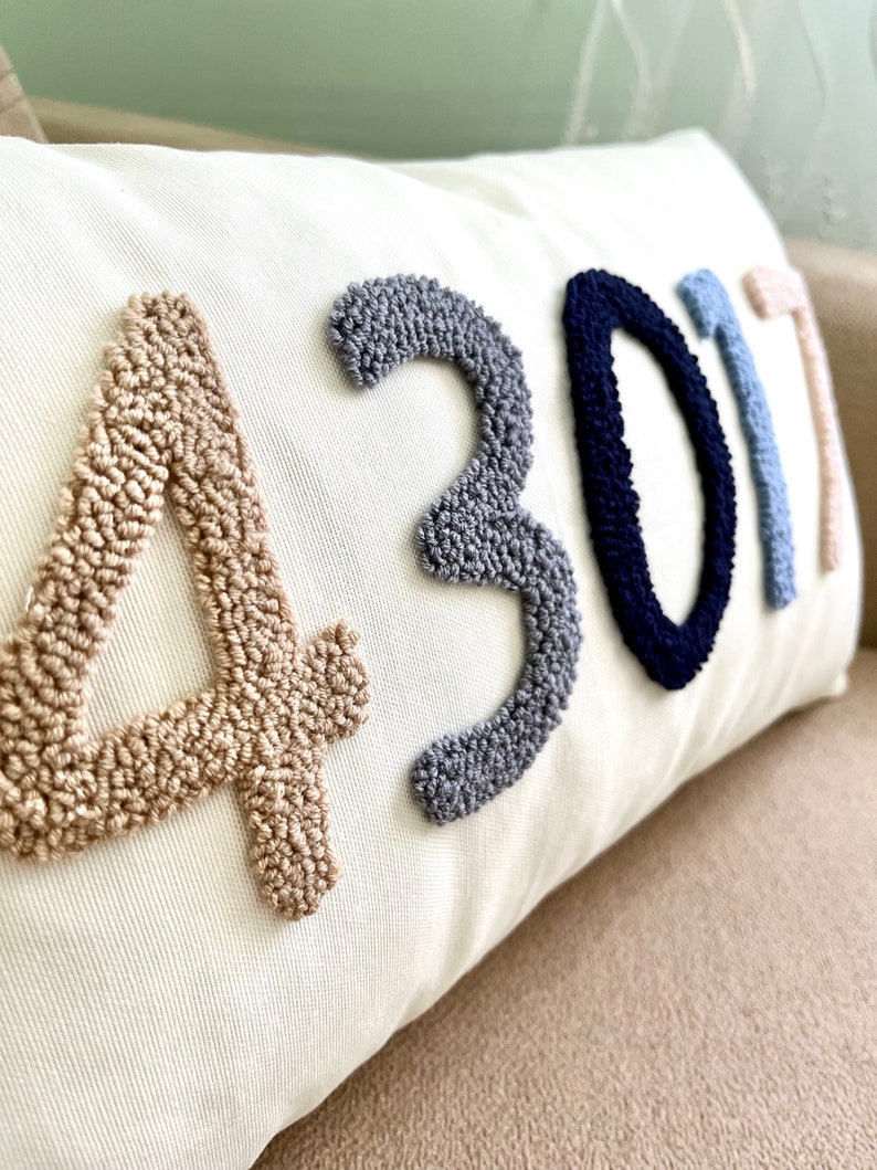 Op de afbeelding: Een wit kussen met de cijfers 4301 geborduurd in beige, grijs, donkerblauw en lichtblauw garen.