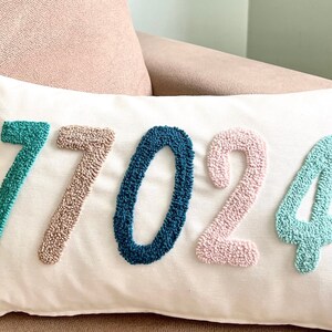 May include: A white pillow with the numbers 77024 embroidered in turquoise, beige, teal, pink, and light turquoise.