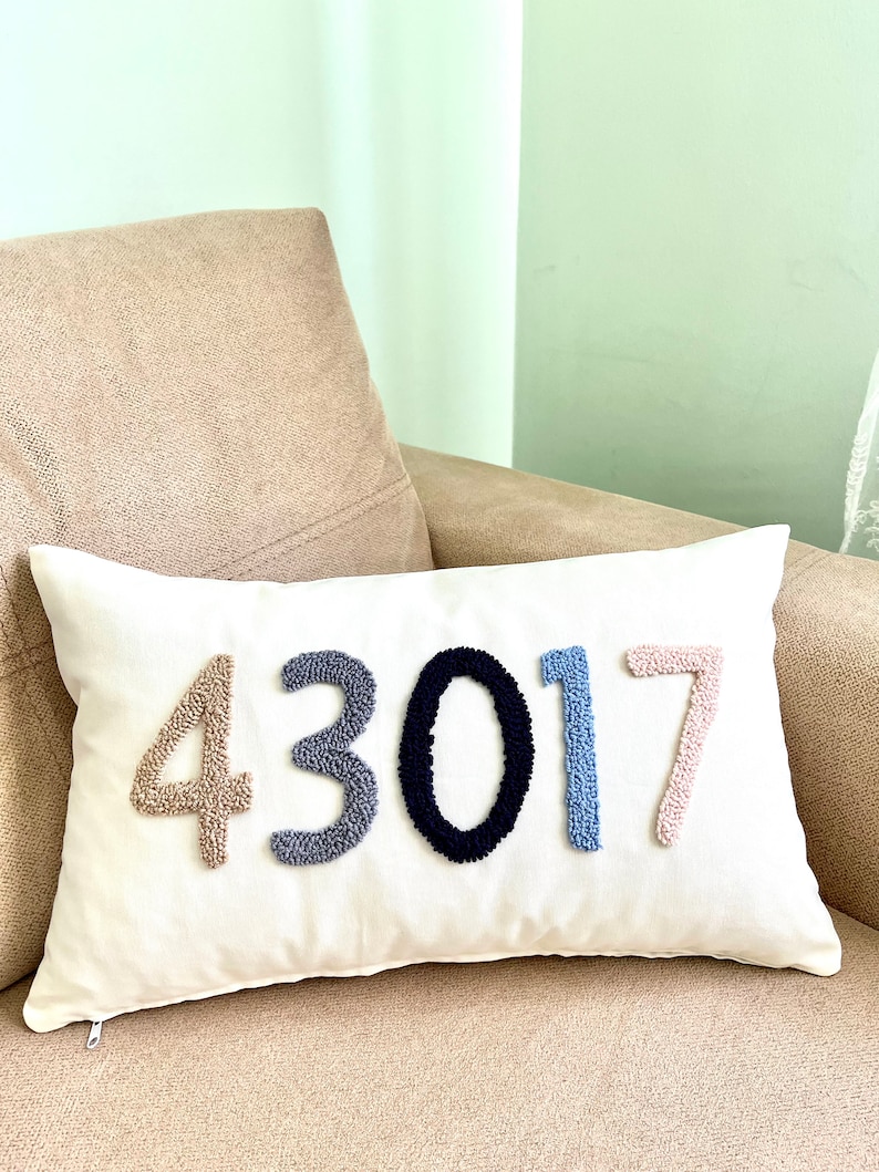 May include: A white pillow with the number 43017 embroidered in colorful yarn. The numbers are in different colors: beige, gray, black, blue, and pink.