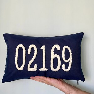 Op de afbeelding: Een donkerblauw kussen met witte, pluche letters die "02169" lezen.