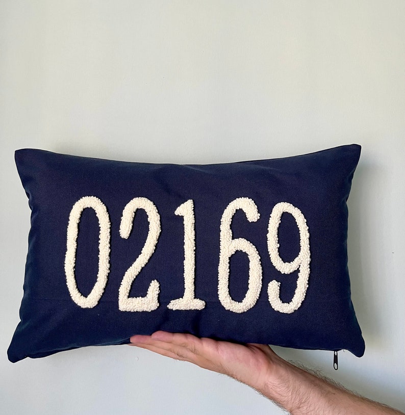 Op de afbeelding: Een donkerblauw kussen met witte, pluche letters die "02169" lezen.