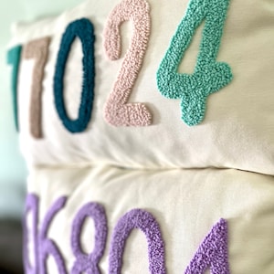 May include: Two white pillow covers with embroidered numbers. The top pillow has the numbers 17024 in teal, pink, and light green. The bottom pillow has the numbers 06804 in purple.