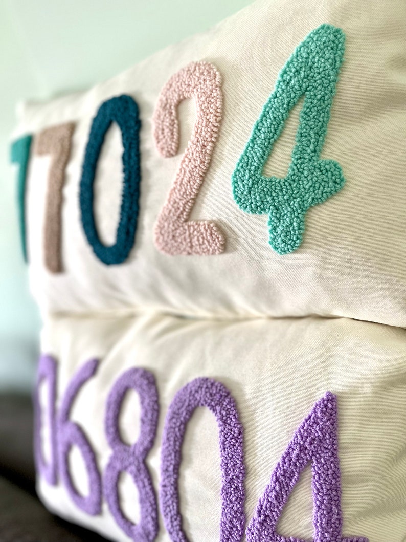 May include: Two white pillow covers with embroidered numbers. The top pillow has the numbers 17024 in teal, pink, and light green. The bottom pillow has the numbers 06804 in purple.