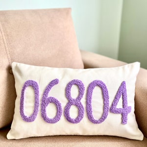 Op de afbeelding: Een wit kussen met het nummer 06804 geborduurd in paars. Het kussen ligt op een lichtbruine bank.