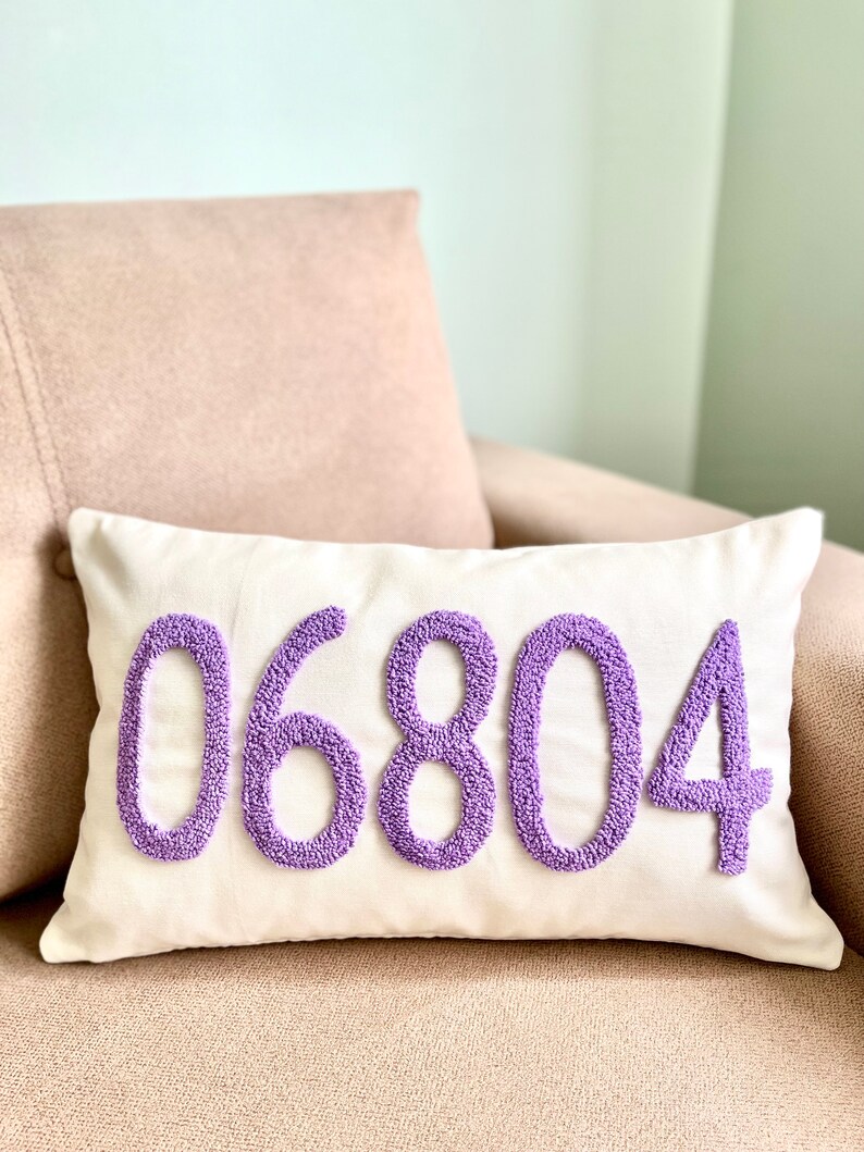 Op de afbeelding: Een wit kussen met het nummer 06804 geborduurd in paars. Het kussen ligt op een lichtbruine bank.