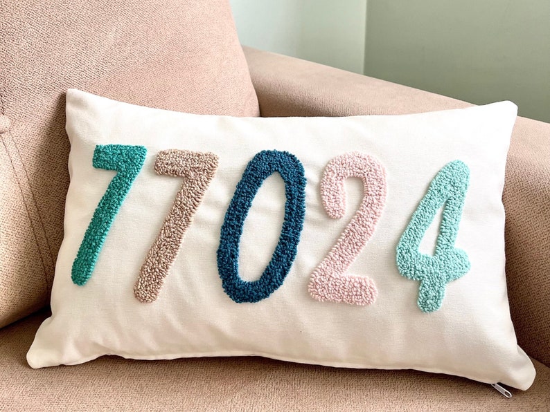Op de afbeelding: Een wit kussen met de cijfers 77024 geborduurd in verschillende kleuren. De cijfers zijn in een regenboogkleurenschema, beginnend met teal, dan beige, dan donkerblauw, dan roze en eindigend met licht teal.