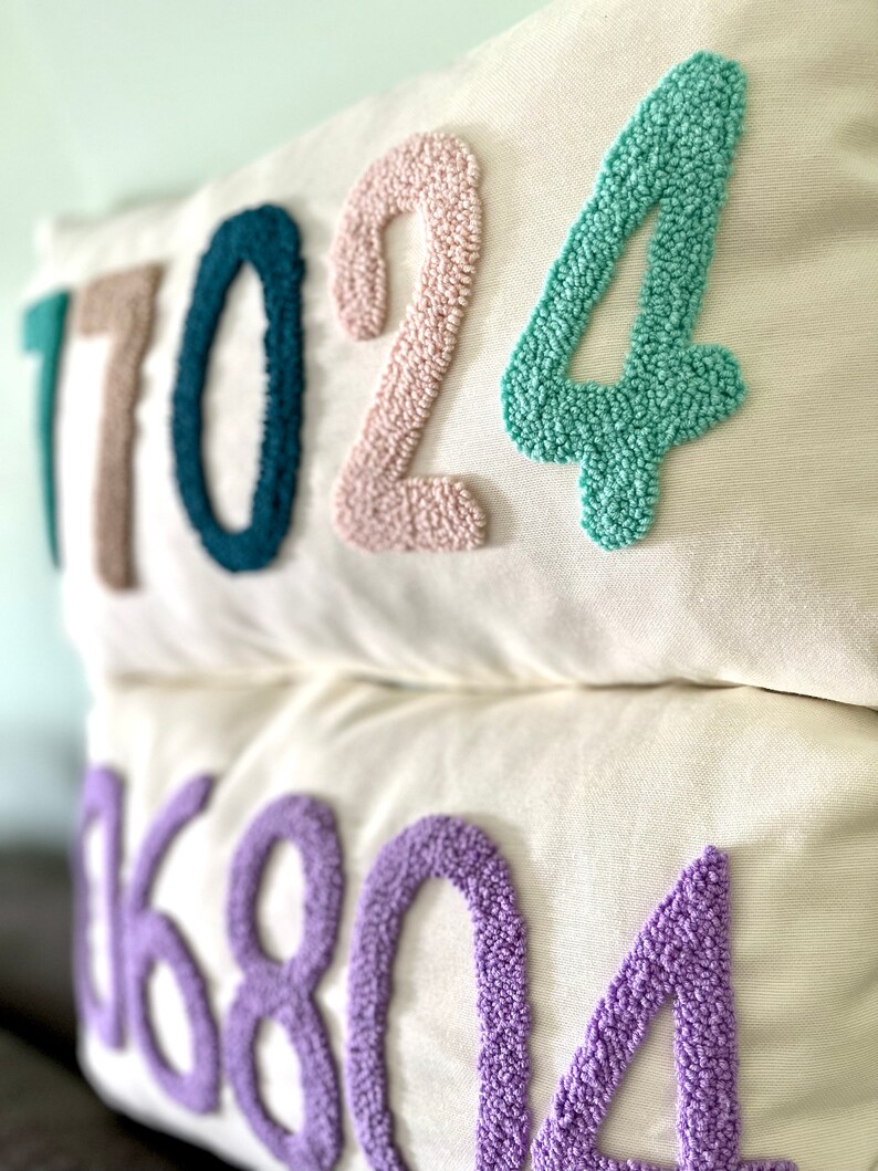 Op de afbeelding: Twee witte kussenhoezen met geborduurde nummers. Het bovenste kussen heeft de nummers 17024 in teal, roze en lichtgroen. Het onderste kussen heeft de nummers 06804 in paars.