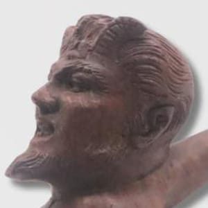 May include: A brown wooden tobacco pipe carved into the shape of a horned figure's head. The carving features detailed facial expressions, including a pointed beard, textured hair, and visible horns on the forehead, connected to a long curved stem.