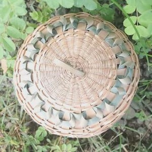 May include: A round, woven basket with a lid featuring a central handle. The basket is constructed from light tan natural fibers and accented with a decorative border of sage green ribbon woven through the outer edge of the circular lid.