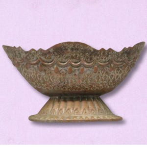 May include: A bronze-colored decorative bowl with a scalloped rim and a pedestal base. The bowl features intricate embossed patterns and a detailed design. The bowl is likely made of metal and is suitable for display or decorative purposes.