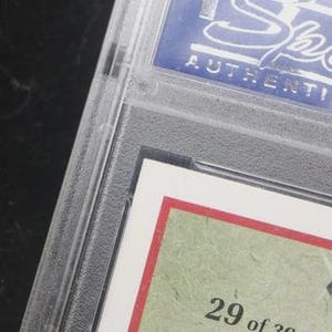 May include: A sports trading card encased in a clear plastic holder. The card has a red border and a green background with the number "29 of 30" visible. The top of the holder has the word "Sports" and "AUTHENTICATED" in white text on a blue background.