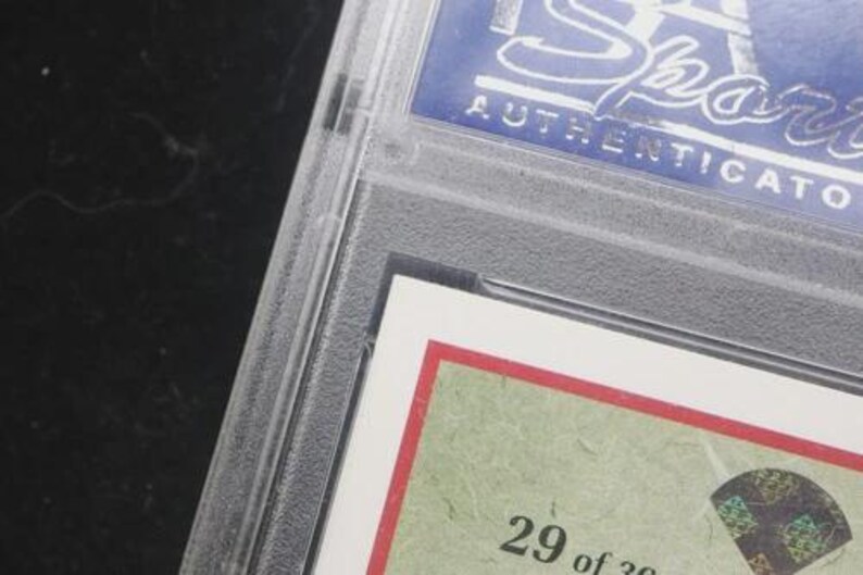 May include: A sports trading card encased in a clear plastic holder. The card has a red border and a green background with the number "29 of 30" visible. The top of the holder has the word "Sports" and "AUTHENTICATED" in white text on a blue background.
