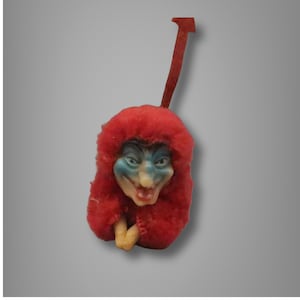 May include: A vintage doll ornament with a red fuzzy hood and a face with blue and beige paint. The doll has a red pull tab.