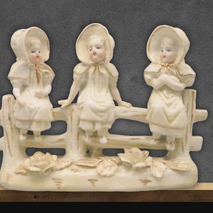 May include: A cream-colored porcelain figurine depicting three young girls in bonnets and dresses, seated on a fence. The figurine has a vintage aesthetic, with floral accents at the base. The girls are posed in different positions.