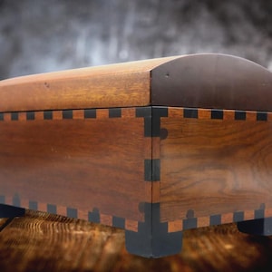 May include: A wooden chest with a curved lid. The chest is made of dark brown wood with black accents at the corners and base. The lid is a darker shade of brown. The chest sits on four small, black feet.