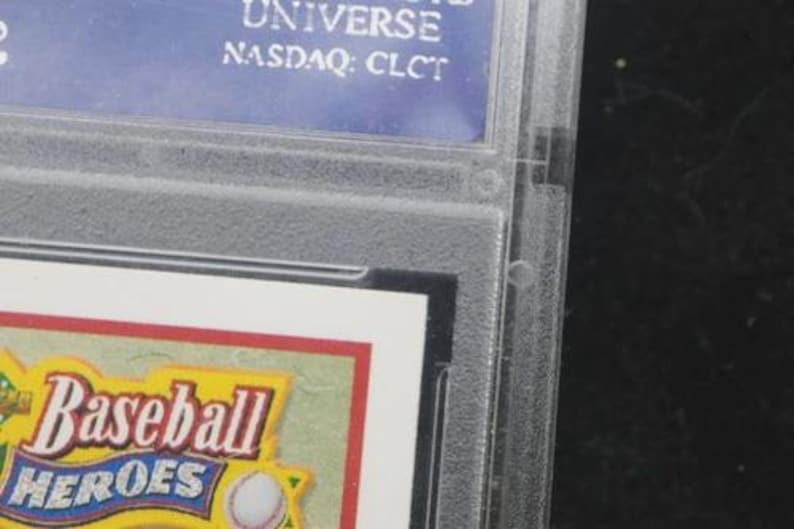May include: A close-up of a baseball card encased in a clear plastic holder. The card features the words "Baseball Heroes" in a yellow and red design. The top of the holder has a blue label with white text, including "UNIVERSE" and "NASDAQ: CLCT".