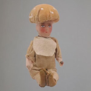 May include: A doll with a pale face, rosy cheeks, and blue eyes, wearing a tan fabric outfit with a light beige bib. The doll has a large, rounded, tan mushroom-shaped hat covering its head, featuring subtle white spots on the top surface.