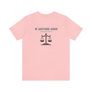 Op de afbeelding: Roze T-shirt met de tekst "IF ANYONE ASKS I plead the FIFTH" en een zwarte weegschaal van de rechtvaardigheid.