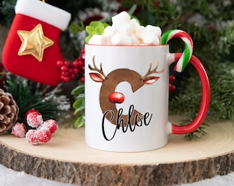 May include: White ceramic mug with a red handle and a candy cane attached. The mug has a reindeer illustration with the letter "C" and the name "Chloe" written in black cursive.