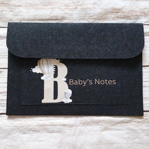 May include: A dark grey felt envelope case with a flap closure. It features a decorative letter "B" with a hot air balloon design and the text "Baby's Notes" in light brown. The case is designed for baby-related items, made of felt.