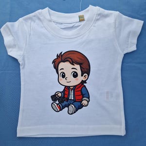 May include: White short-sleeved t-shirt with a cartoon illustration of a character with brown hair, a red vest, and blue jeans. The character is holding sunglasses. The shirt is designed for children.