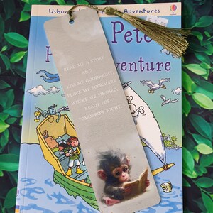 May include: A beige bookmark with a monkey reading a book. The bookmark has the text "Read me a story and kiss me goodnight, place my bookmark where we finished, ready for tomorrow night." The bookmark is sitting on top of a book titled "Peter's Adventures".