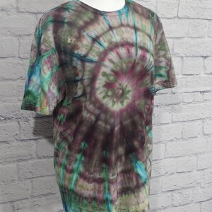 May include: A short-sleeved t-shirt with a spiral tie-dye pattern. The shirt features a blend of colors, including shades of purple, green, and blue. The tie-dye design radiates from the center of the shirt, creating a unique and colorful look.