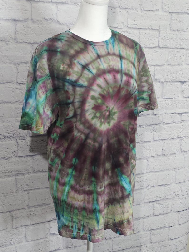 May include: A short-sleeved t-shirt with a spiral tie-dye pattern. The shirt features a blend of colors, including shades of purple, green, and blue. The tie-dye design radiates from the center of the shirt, creating a unique and colorful look.