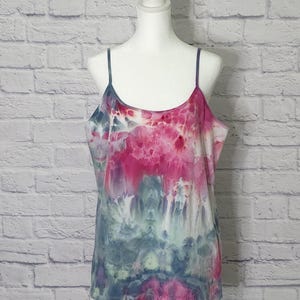 May include: A tie-dye camisole in shades of pink, blue, and white, displayed on a mannequin. The top features thin spaghetti straps and a scoop neckline. The fabric has a watercolor-like effect, with the colors blending together.