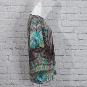 May include: A tie-dye t-shirt in shades of teal, purple, and green is displayed on a white mannequin. The short-sleeved shirt has a round neckline and a unique, swirling pattern. The background is a white brick wall.
