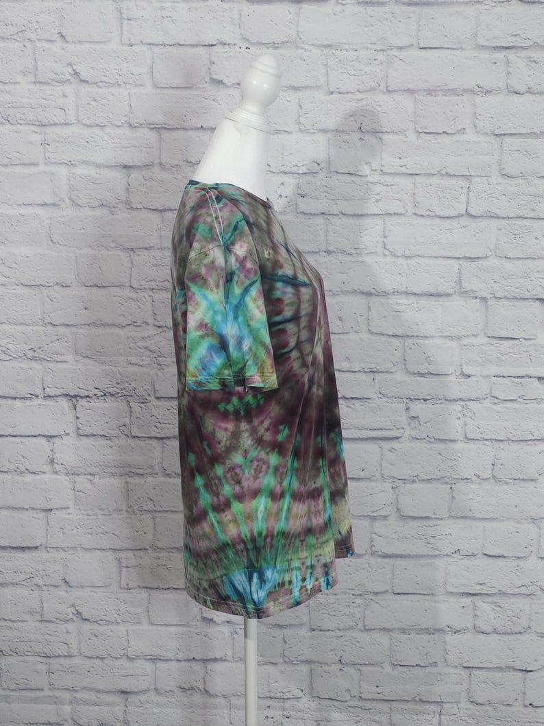 May include: A tie-dye t-shirt in shades of teal, purple, and green is displayed on a white mannequin. The short-sleeved shirt has a round neckline and a unique, swirling pattern. The background is a white brick wall.