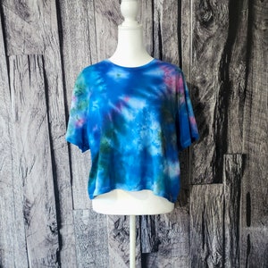 May include: A blue, green, and pink tie-dye cropped t-shirt on a white mannequin against a wood background.