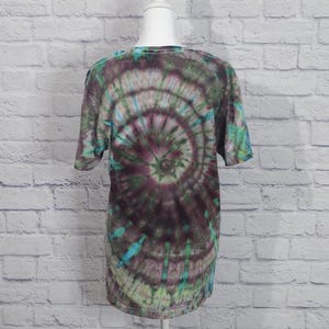 May include: A short-sleeved tie-dye t-shirt in shades of green, teal, and purple. The shirt features a spiral pattern radiating from the center. The shirt is displayed on a white mannequin head and torso against a white brick wall.