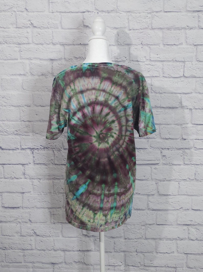 May include: A short-sleeved tie-dye t-shirt in shades of green, teal, and purple. The shirt features a spiral pattern radiating from the center. The shirt is displayed on a white mannequin head and torso against a white brick wall.