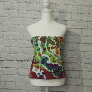 May include: A strapless top with a vibrant tie-dye pattern in shades of red, green, yellow, and blue. The top is displayed on a white mannequin against a white brick wall background. The garment has a fitted silhouette.