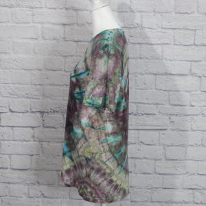 May include: A tie-dye t-shirt in shades of purple, teal, and green. The short-sleeved shirt is displayed on a white mannequin. The fabric has a swirling pattern, with the colors blending together. The shirt has a round neckline.