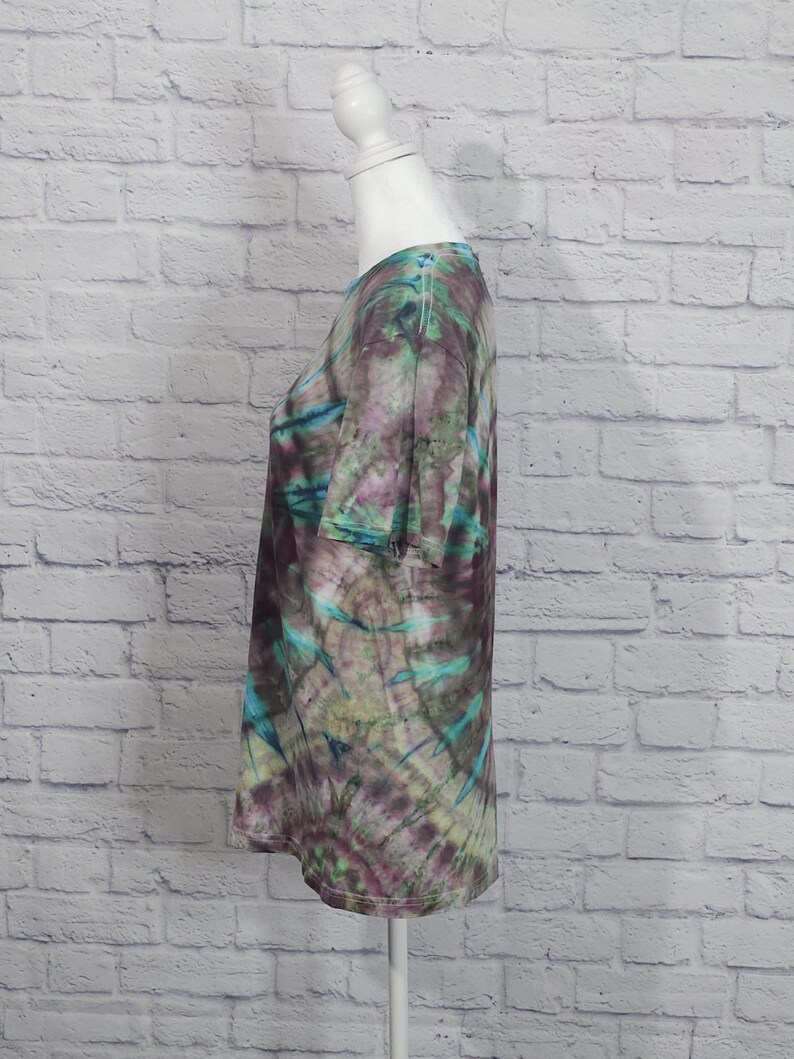 May include: A tie-dye t-shirt in shades of purple, teal, and green. The short-sleeved shirt is displayed on a white mannequin. The fabric has a swirling pattern, with the colors blending together. The shirt has a round neckline.