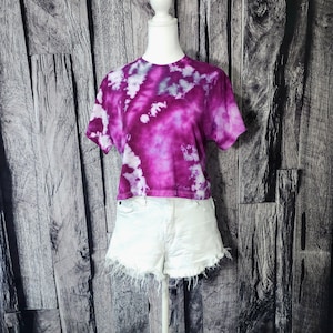 May include: A tie-dye cropped t-shirt in shades of purple and white. The shirt is on a white mannequin and has short sleeves.