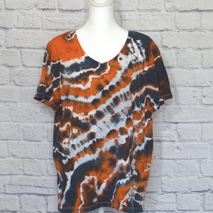 May include: A short-sleeved, tie-dye V-neck t-shirt in orange, white, and dark blue. The shirt is displayed on a white mannequin against a white brick wall. The tie-dye pattern has a striped design.