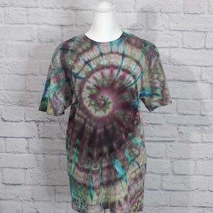 May include: A short-sleeved tie-dye t-shirt in shades of purple, green, and blue. The design features a spiral pattern radiating from the center. The shirt is displayed on a white mannequin against a white brick wall.