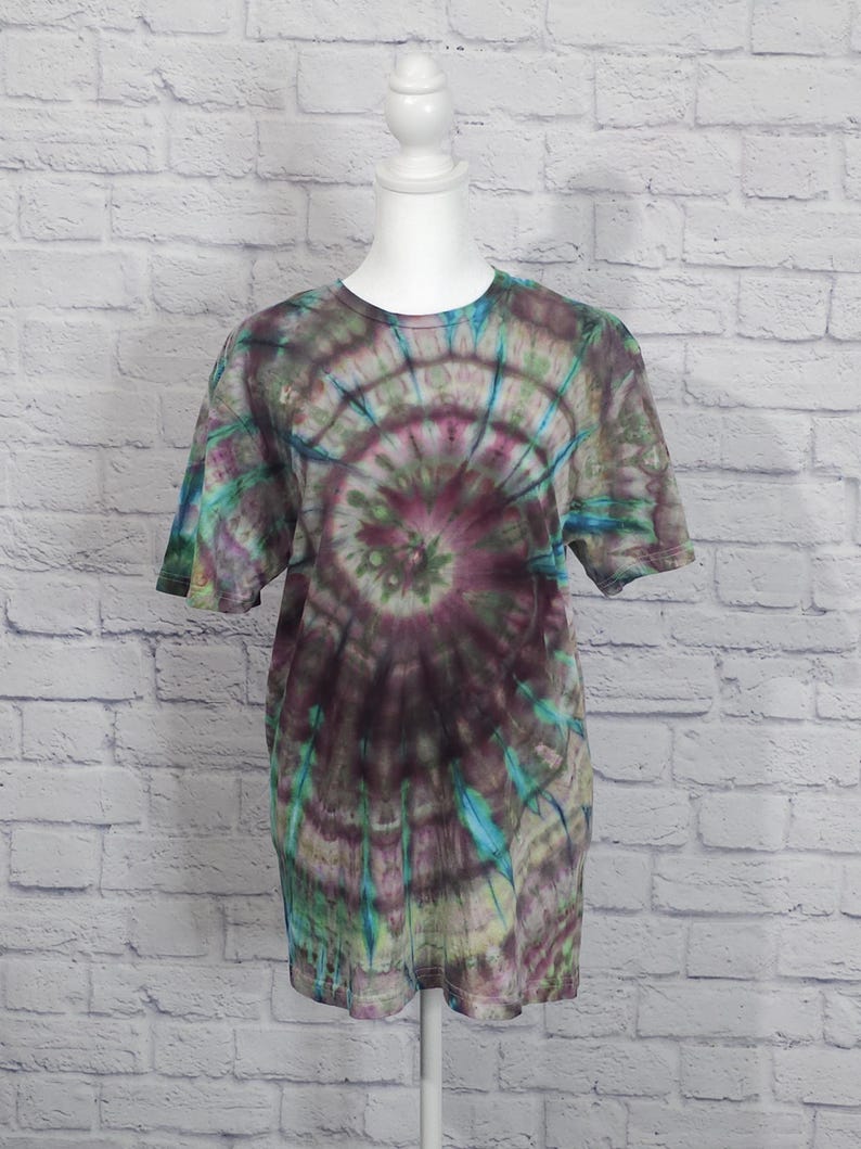 May include: A short-sleeved tie-dye t-shirt in shades of purple, green, and blue. The design features a spiral pattern radiating from the center. The shirt is displayed on a white mannequin against a white brick wall.