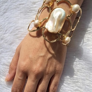 May include: Gold-toned cuff bracelet with a large, iridescent pearl center and smaller pearls. The bracelet has an openwork design and is labeled "Elegant Pearl Cuff" and "Handmade Luxury Bracelet".