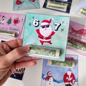 May include: A hand holds a Christmas-themed gift card featuring Santa Claus wearing sunglasses and holding the numbers 6 and 7. The card is teal with a dollar bill in a clear plastic sleeve. Other cards with festive designs are in the background.