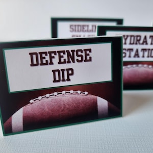 May include: Cards with football-themed designs. The front card reads "DEFENSE DIP" in brown letters on a white background, framed in green. A football graphic is at the bottom. Other cards say "SIDELINE" and "HYDRATION STATION".