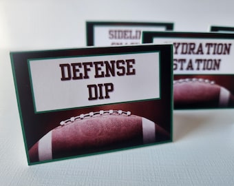 May include: Cards with football-themed designs. The front card reads "DEFENSE DIP" in brown letters on a white background, framed in green. A football graphic is at the bottom. Other cards say "SIDELINE" and "HYDRATION STATION".