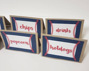 May include: Set of five brown cardstock table signs with blue and red baseball-themed borders. The signs read "chips", "popcorn", "drinks", "hotdogs", and an empty sign.