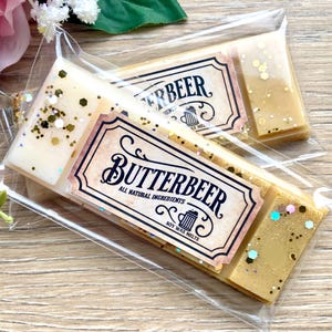 May include: Two soy wax melts in clear packaging with gold glitter. The wax melts are labelled "Butterbeer" with the text "All Natural Ingredients" and "Soy Wax Melts".