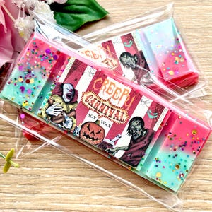May include: Two rectangular soy wax melts in clear plastic packaging. The wax is pink and light blue with multicolored glitter. A label features red and white stripes, a clown, a pumpkin, a zombie, and the text Creepy Carnival Soy Wax.