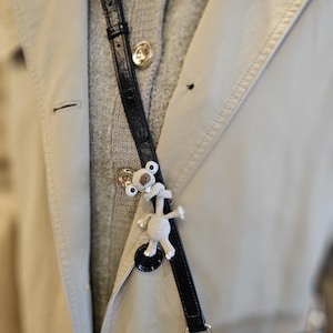 Pode incluir: Uma alça de bolsa de couro preta com um pequeno pingente de coala de crochê. O coala é branco e bege, com olhos pretos e um nariz pequeno. A alça está presa a um trench coat bege.