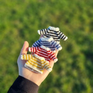 May include: Four miniature knitted sweaters in a stack. The sweaters are striped in yellow and white, red and white, blue and white, and black and white.